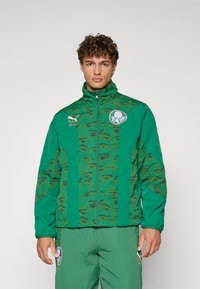 Grüne Zip-Jacke mit einem Camouflage-Muster, das Gesichter zeigt, mit einem Markenlogo-Patch, hohem Kragen und elastischen Bündchen. Das Material scheint leicht und strukturiert zu sein.