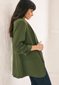 Blazer verde oliva con hombros estructurados, un corte relajado y mangas dobladas, que cuenta con un bolsillo único y una textura de tela suave.