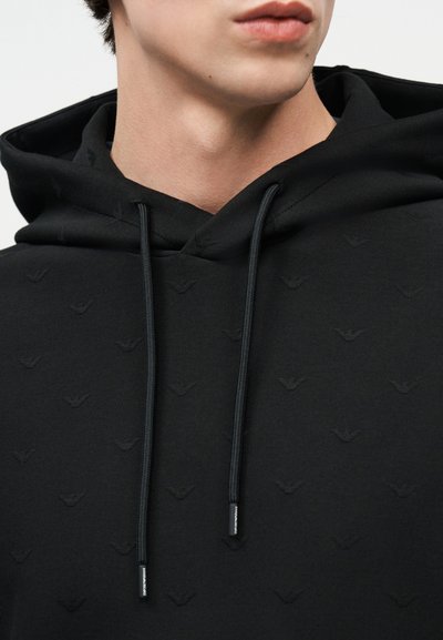 Νεαρό άτομο που φοράει μαύρο φούτερ με κουκούλα, με διακριτικά ανάγλυφα σχέδια και μαύρα κορδόνια, ορατό από το λαιμό μέχρι το στήθος.