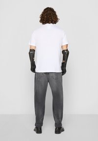 Person med krøllet hår iført en hvid skjorte, grå jeans, sorte sko og sorte proteser til underarmene, som vender væk fra kameraet på en ensfarvet baggrund.