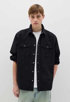 Camicia in denim nero con due tasche sul petto, bottoni a pressione e maniche arrotolate, indossata sopra una maglietta bianca e jeans blu.