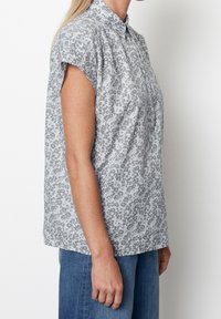 Harmaa lyhythihainen kauluksellinen nappipaita, jossa on kukkakuvio ja hillitty tekstuuri, yhdistetty sinisiin farkkuihin.