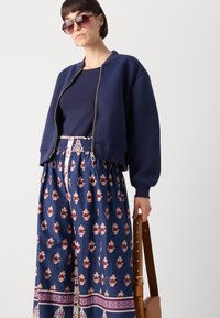 Veste bomber cropped bleu marine sur une chemise bleu marine, associée à une longue jupe à motifs floraux en bleu foncé et bordeaux. Sac bandoulière marron.