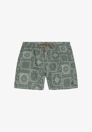 Tânăr stând, purtând o cămașă bej cu nasturi, pantaloni scurți verzi cu model, papuci de plajă negri, ținând un prosop în dungi într-o mână.