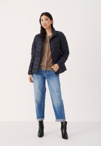 Dunkelblauer Puffer-Jacke mit Kragen und gestepptem Design. Kombiniert mit einem hellbraunen Cardigan und hochgekrempelten blauen Jeans. Schwarze Stiefeletten.