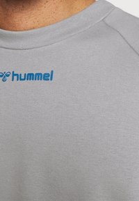T-shirt de sport gris avec un tissu texturé, arborant un logo "hummel" bleu sur le côté gauche de la poitrine. Encolure ronde.