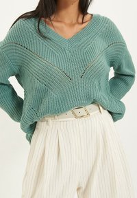 Pull en maille bleu sarcelle avec col en V et motifs texturés, associé à un pantalon plissé à rayures crème et une ceinture de couleur claire.