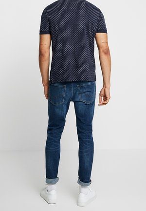Homme portant une chemise bleu foncé à manches courtes à pois, jean bleu retroussé, chaussettes blanches et baskets blanches, vu de dos.