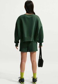 Sweat-shirt oversize vert foncé et short assorti avec une finition texturée. Sac bandoulière noir et chaussures sportives avec des accents néon.
