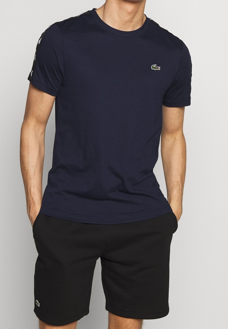Navy blue cotton t-shirt with short sleeves, featuring a small green crocodile logo on the chest and contrasting striped accents on the shoulders.
