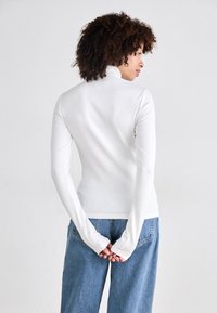 Lange mouwen, witte, getailleerde top met een hoge hals. De stof is glad met een ribbelstructuur. Gecombineerd met losse, blauwe denim jeans.