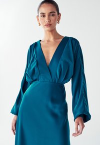 Žena v tyrkysových saténových šatech s dlouhým rukávem a hlubokým výstřihem do V, se zlatými kruhovými náušnicemi, stojící na jednolitém bílém pozadí.