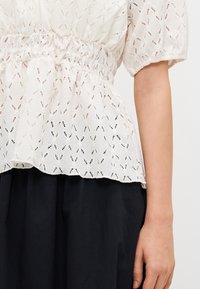 Weiße Bluse mit strukturierter Augenmusterung, elastischem Bund und kurzen Ärmeln, kombiniert mit einem dunklen, voluminösen Rock.