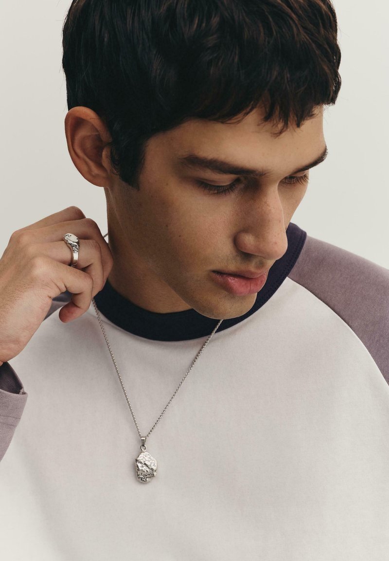 Silberne Halskette mit einem strukturierten Anhänger, getragen über einem weißen und lila Raglan-Shirt; die Hand des Modells ist in der Nähe der Halskette positioniert.