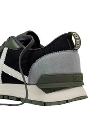 Zapatilla negra y gris con ante texturizado, parte superior de malla, acentos blancos y suela de goma verde. Presenta una lengüeta trasera prominente para facilitar el calzado.