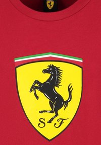Czerwona koszulka z okrągłym dekoltem, z żółtym logo w kształcie tarczy. Logo zawiera czarnego konia w skoku oraz tekst "SF" u podstawy.