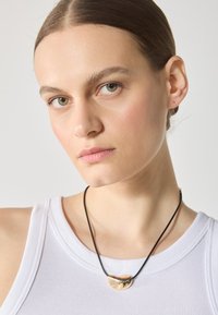 Colier cu pandantiv aurit, având un design neted și curbat, pe un șnur negru. Purtați-l peste o bluză albă fără mâneci, cu textură fagure.