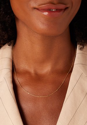 Un collier en chaîne dorée présente un design délicat avec de petits maillons ronds, porté contre une peau bronzée et texturée.