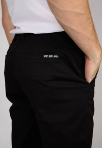 Pantalons en coton noir avec une poche arrière ornée d'un patch bleu représentant trois petites silhouettes d'animaux. Design ajusté avec une texture lisse.