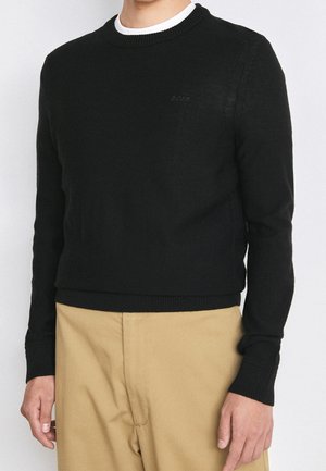 Man draagt een zwarte ronde hals trui met een subtiel "BOSS" logo, gecombineerd met beige broek, zichtbaar van nek tot taille.
