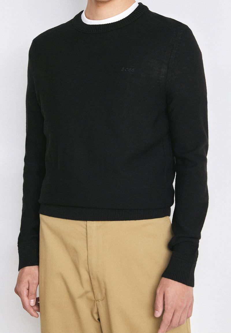 Homme portant un pull à col rond noir avec un logo "BOSS" subtil, associé à un pantalon beige, photographié du cou à la taille.