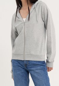 Sudadera gris con cremallera hecha de tejido suave, con capucha, bolsillos frontales y puños acanalados. Llevada sobre una camisa blanca y combinada con vaqueros azules.