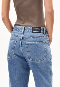 Närbild av en person som bär ljusblå denimjeans med stora bakfickor och en svart Arnedangels-logotyp, tillsammans med en brun ärmlös topp.