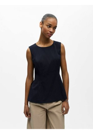 Femme portant un haut sans manches à fines rayures foncées et un pantalon beige, debout contre un fond blanc uni avec une posture détendue.