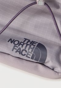 Κοντινό πλάνο ενός γκρι τμήματος υφάσματος με μαύρο λογότυπο The North Face και μοβ θηλιά κορδονιού σε υφή υλικού.