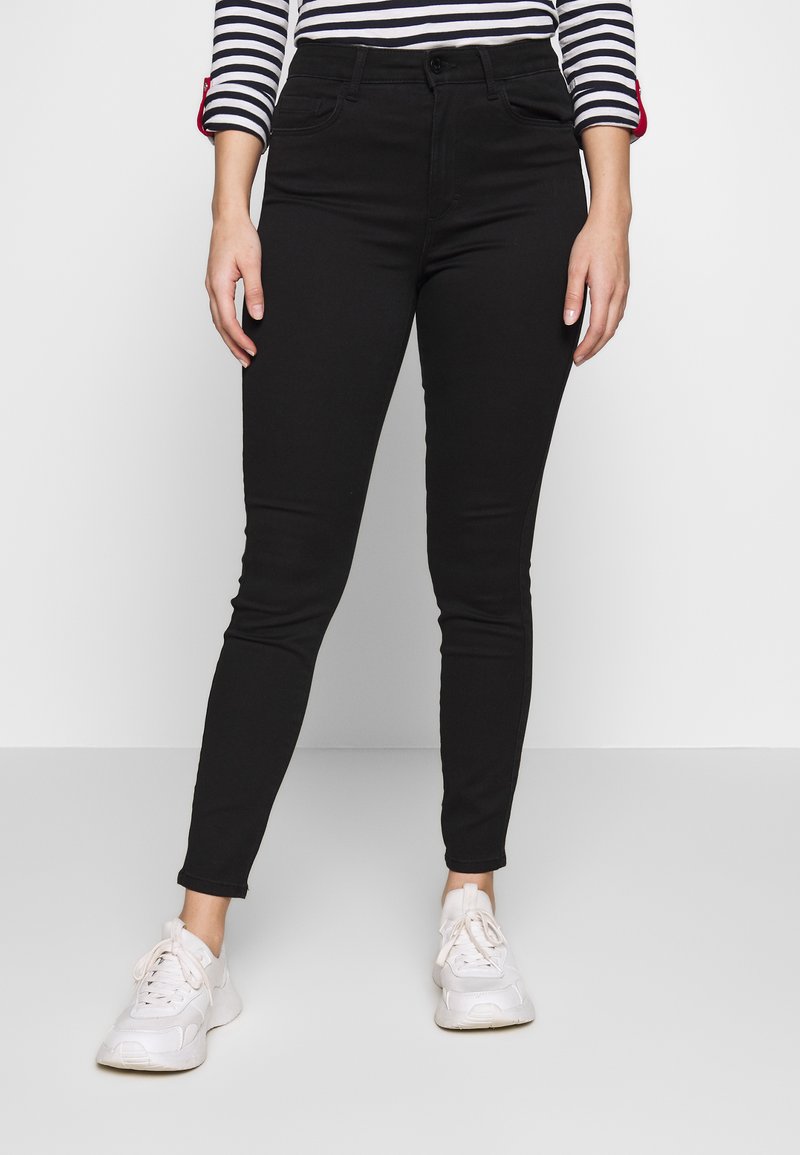 Jeans slim fit pretos com uma textura suave, apresentando uma cintura de altura média, bolsos frontais e um fecho simples com botão. Combinados com sapatilhas brancas.