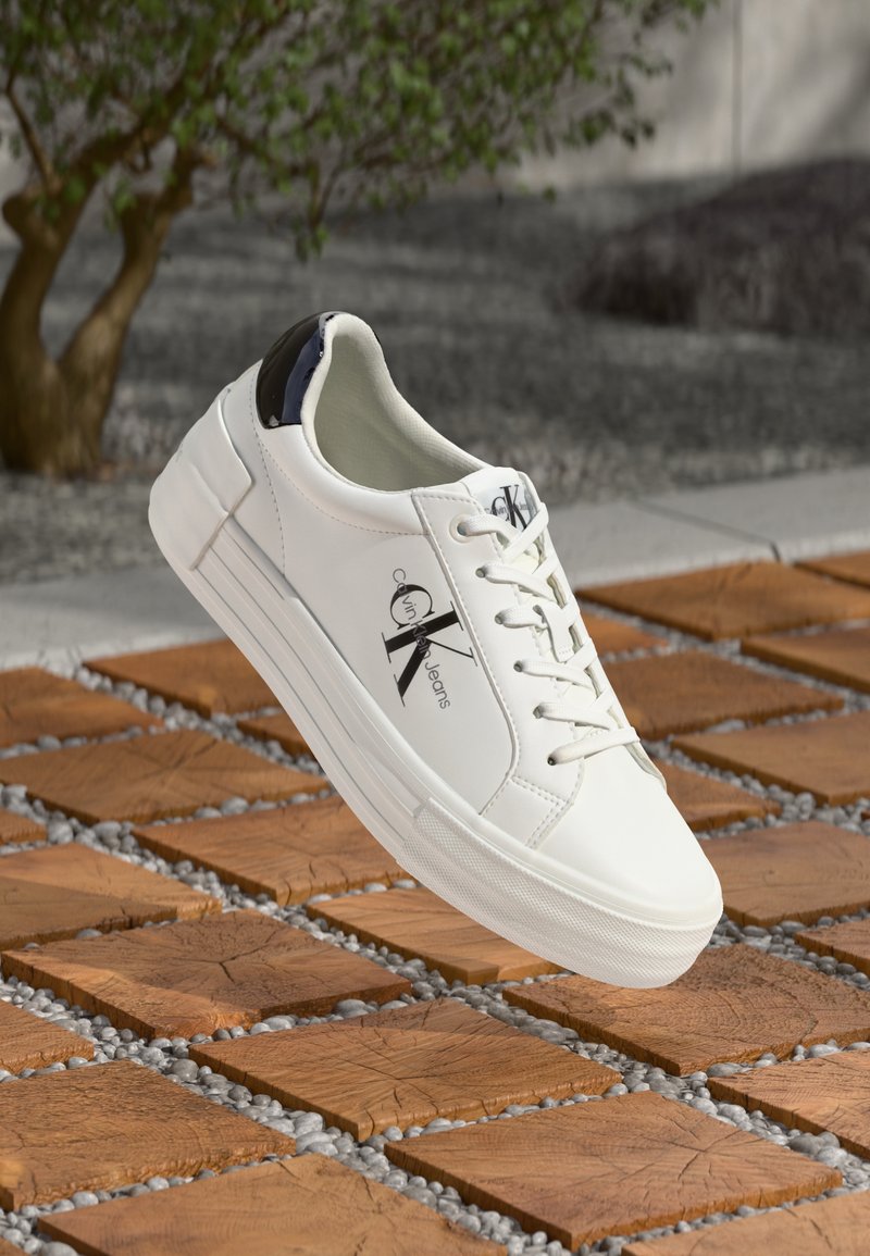 White Calvin Klein sneaker with black heel detail floating above wooden blocks and small gray stones outdoors.