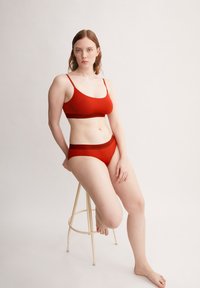 Soutien-gorge rouge et culotte assortie en tissu lisse, avec des bretelles fines et une ceinture avec le logo "ARMEDANGELS". Modèle assis sur un tabouret.