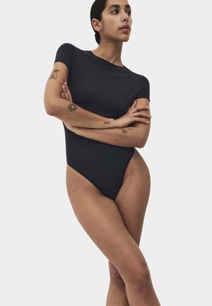 Kvinde står med armene krydset iført en tætsiddende sort bodysuit med korte ærmer, der viser tatoveringer på hendes arme mod en enkel baggrund.