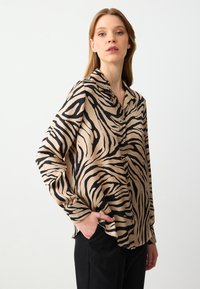 Beige shirt met zwarte zebraprinten, met een kraag, lange mouwen en knopen aan de voorkant, tegen een effen witte achtergrond.