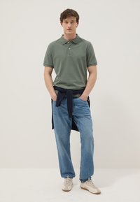 Joven de pie, vistiendo una camiseta polo verde, vaqueros azules, zapatillas blancas y un suéter oscuro atado alrededor de su cintura contra un fondo liso.