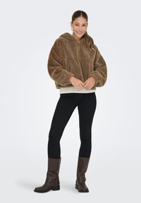 Bruine fleecetop met capuchon, half-rits aan de voorkant, brede mouwen en een relaxte pasvorm, gecombineerd met zwarte leggings en bruine kniehoge laarzen.