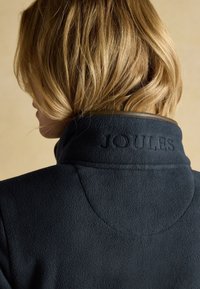Persona bionda vista da dietro che indossa una giacca in pile blu scuro con un colletto alto ricamato con la parola "JOULES."