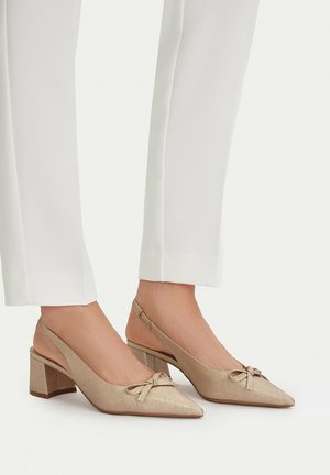 Pies con zapatos de tacón tipo slingback de punta beige, con tacones gruesos y lazos decorativos, combinados con pantalones blancos hasta el tobillo.