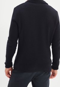 Jersey de punto azul marino oscuro con un patrón texturizado, mangas largas y cuello tipo chal. El dobladillo es ligeramente más largo en la parte trasera.