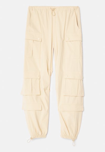 Beige cargo pants with an elastic waist, adjustable drawstring, multiple large pockets, and elastic cuffs at the ankles. Soft fabric texture.