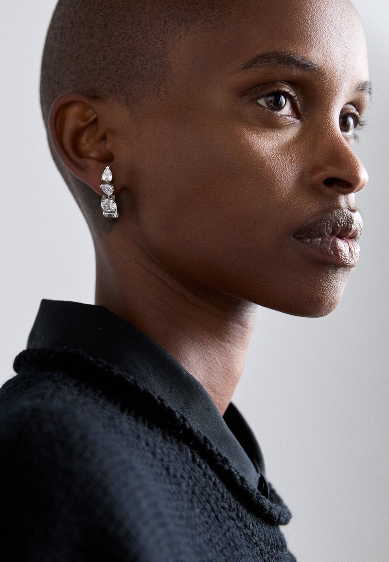 Λαμπερά ασημένια σκουλαρίκια δήλωσης με τρεις γωνιώδεις, περφορέ πέτρες σε διάφορα μεγέθη. Το μοντέλο φοράει ένα μαύρο υφασμάτινο πουλόβερ με γιακά.