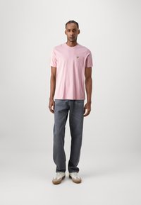Lyserød bomulds T-shirt med rund hals og lille gult logo på brystet, parret med grå denimjeans og hvide sneakers.
