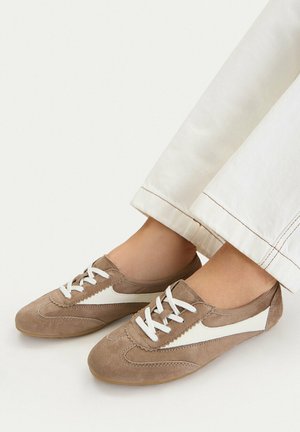 Scarpe casual marroni in camoscio con lacci e strisce laterali bianche, indossate con pantaloni chiari.