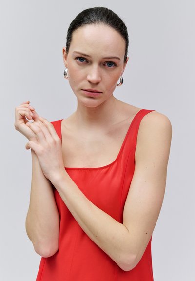 Femme aux cheveux foncés tirés en arrière, portant des boucles d'oreilles argentées en forme de créoles et un haut rouge sans manches, tenant ses mains près de son visage sur un fond uni.