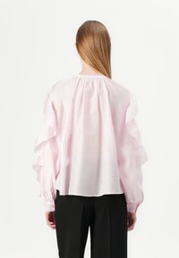 Blusa rosa chiaro con maniche a volant e collo arricciato. Realizzata in tessuto morbido, abbinata a pantaloni neri su misura, vista da dietro.
