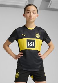 La camiseta de fútbol negra y amarilla presenta un diseño a bloques de color con un gran logo negro, mangas cortas y el emblema de BVB 09 en el lado izquierdo del pecho.