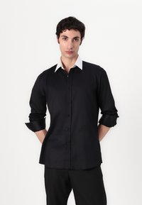 Svart langermet skjorte med hvit krage, knappestolpe, glatt tekstur og brodert detalj nederst til venstre.