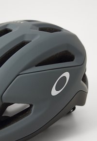 Grauer Fahrradhelm mit elegantem Design, mehreren Belüftungsöffnungen und einem auffälligen weißen Logo an der Seite. Glatte matte Oberfläche.