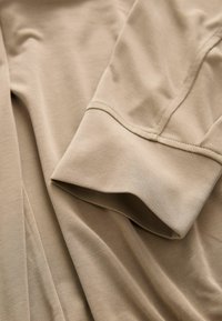 Gros plan sur un tissu beige montrant une manche longue pliée avec une couture visible et un détail de manchette.