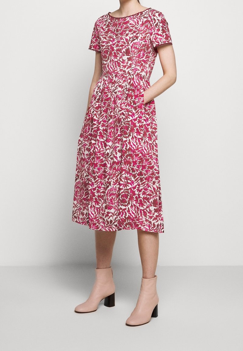 Femme portant une robe midi à manches courtes à fleurs rose et blanche avec poches et bottines nude à talons blocs.